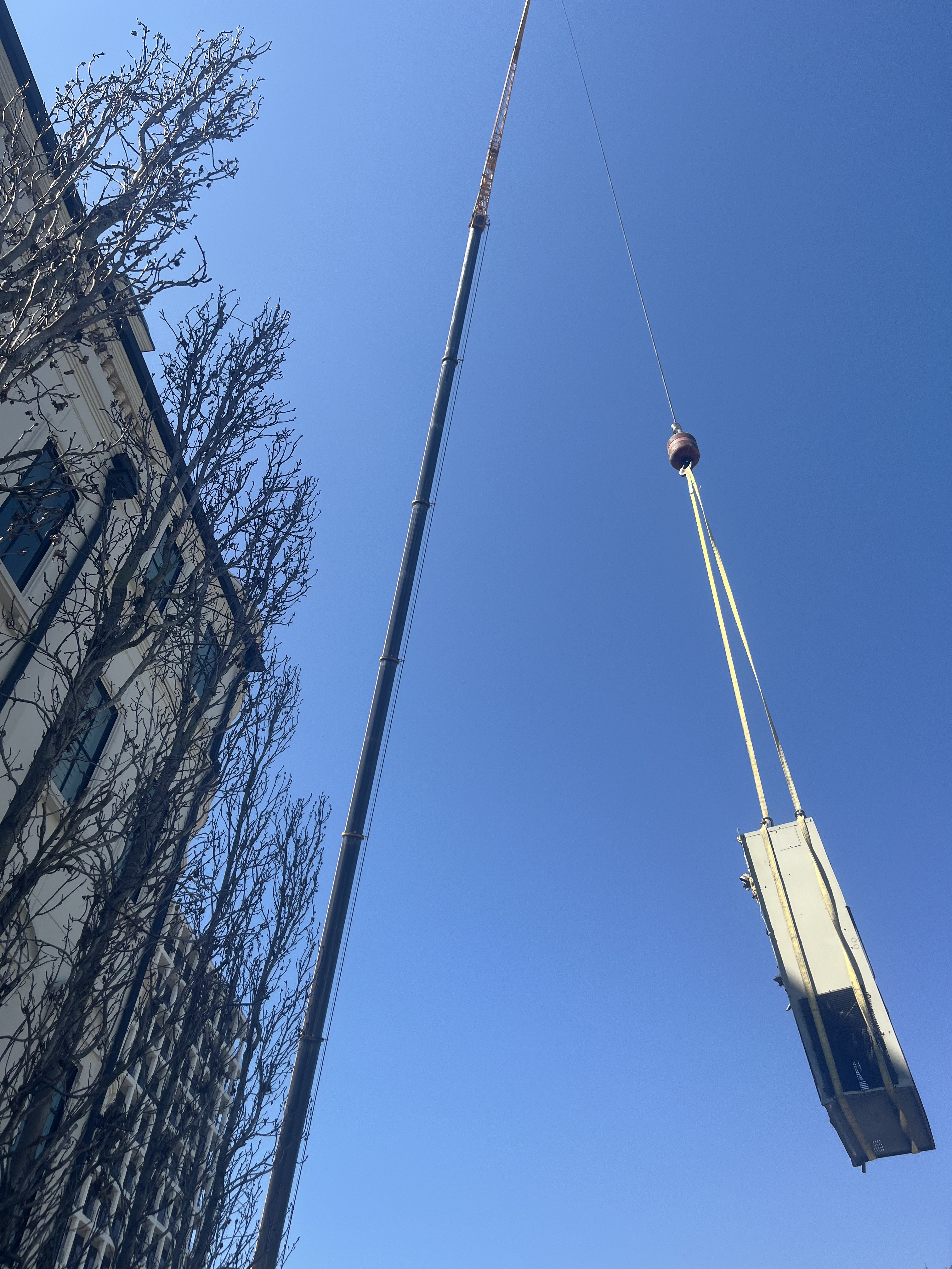 Looking up at crane lifting HVAC unit with yellow straps against blue sky